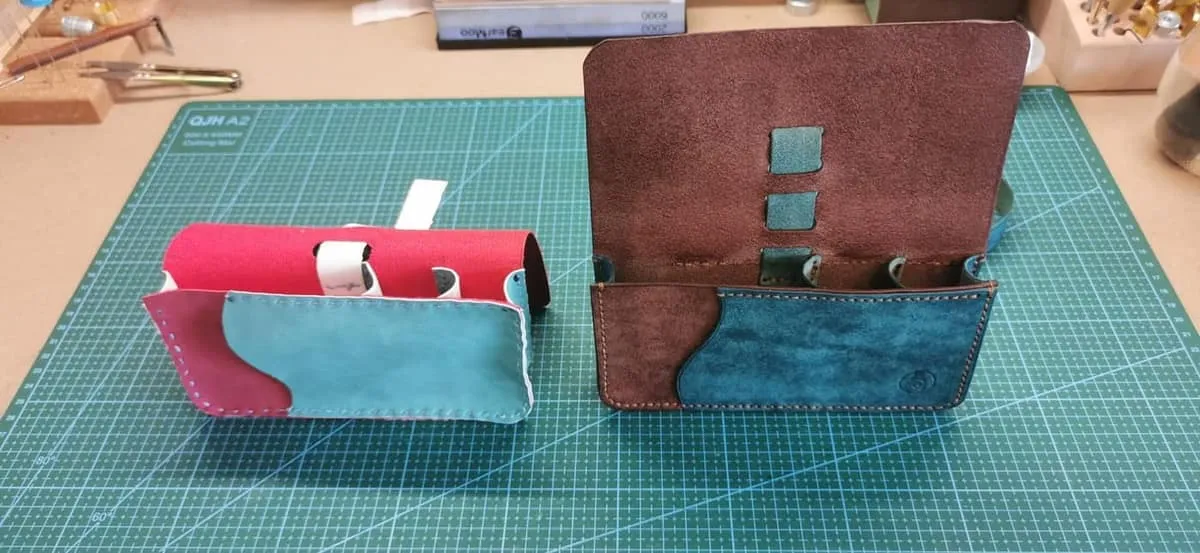 Deux pochettes côte à côte sur un tapis de découpe. À gauche, un prototype grossier en matière rose et turquoise avec des points de couture visibles. À droite, la pochette finie en cuir marron et bleu, ouverte, avec des passants intérieurs et une couture sellier soignée.
